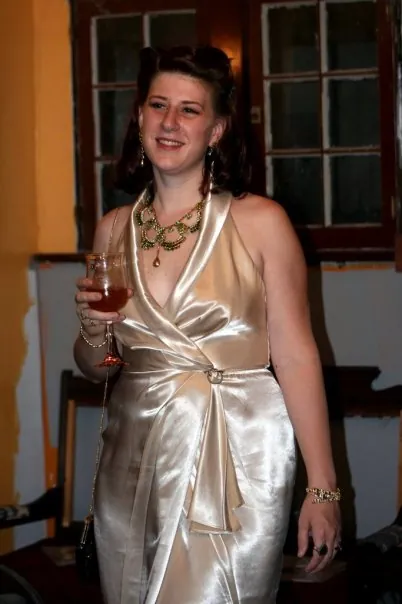 A woman in a champagne satin halter gown holding a glass of wine, wearing a jeweled necklace