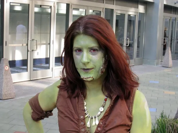 A player with green skin makeup, red eyes, and stitched scar makeup wearing a bone necklace outside a convention center